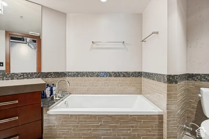 Built-in soaking tub with a tiled surround, wood-finish vanity cabinet, stone countertop, integrated sink, and mosaic glass tile accent detailing