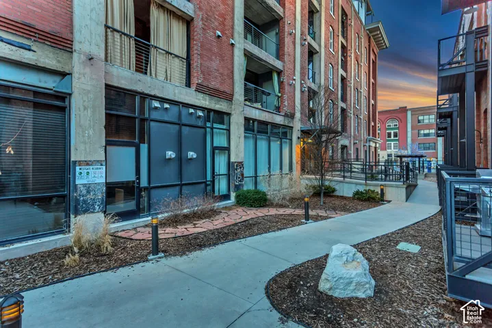 Industrial-style building exterior featuring exposed brick facade, expansive metal-framed window systems, concrete columns, integrated balconies, and a paved walkway with stone hardscaping
