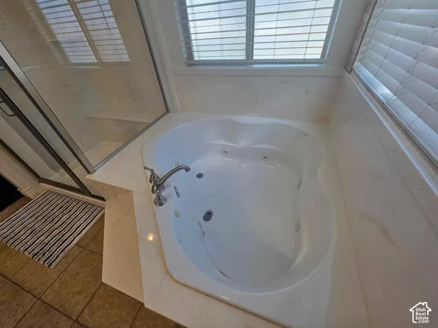 Jetted soaking tub with chrome fixtures, surrounded by light-toned, stone-finish tiling, incorporating a glass-enclosed shower stall and horizontal window blinds
