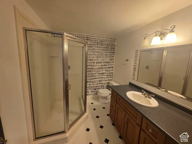 Corner shower stall with chrome-finished frame, wood-toned vanity cabinetry, oversized mirror with integrated multi-bulb lighting, and geometric-patterned tile flooring