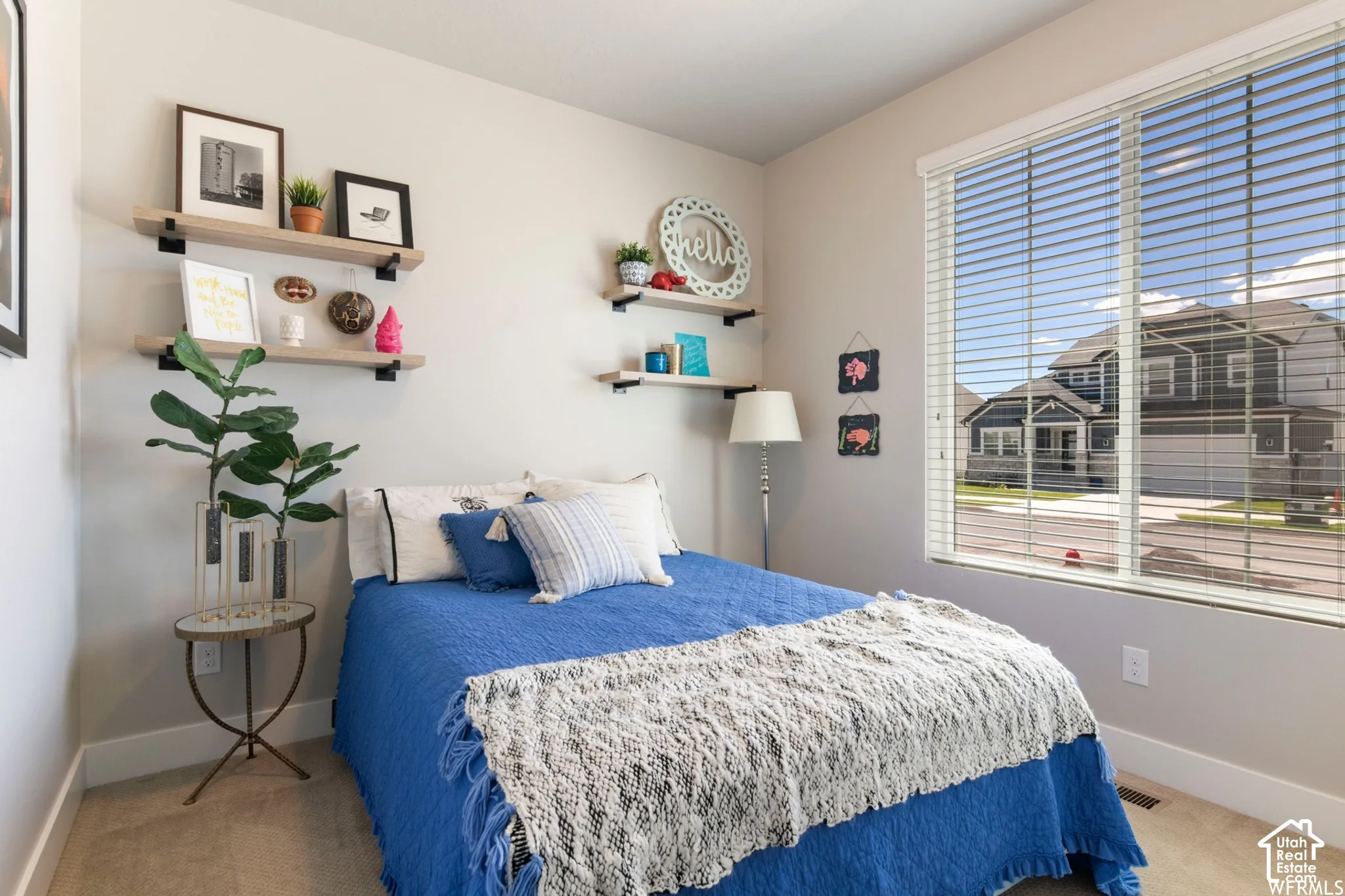 Neutral-toned interior room featuring large window with horizontal blinds, neutral wall-to-wall carpeting, and multiple wall-mounted wood-finish floating shelves with metal brackets