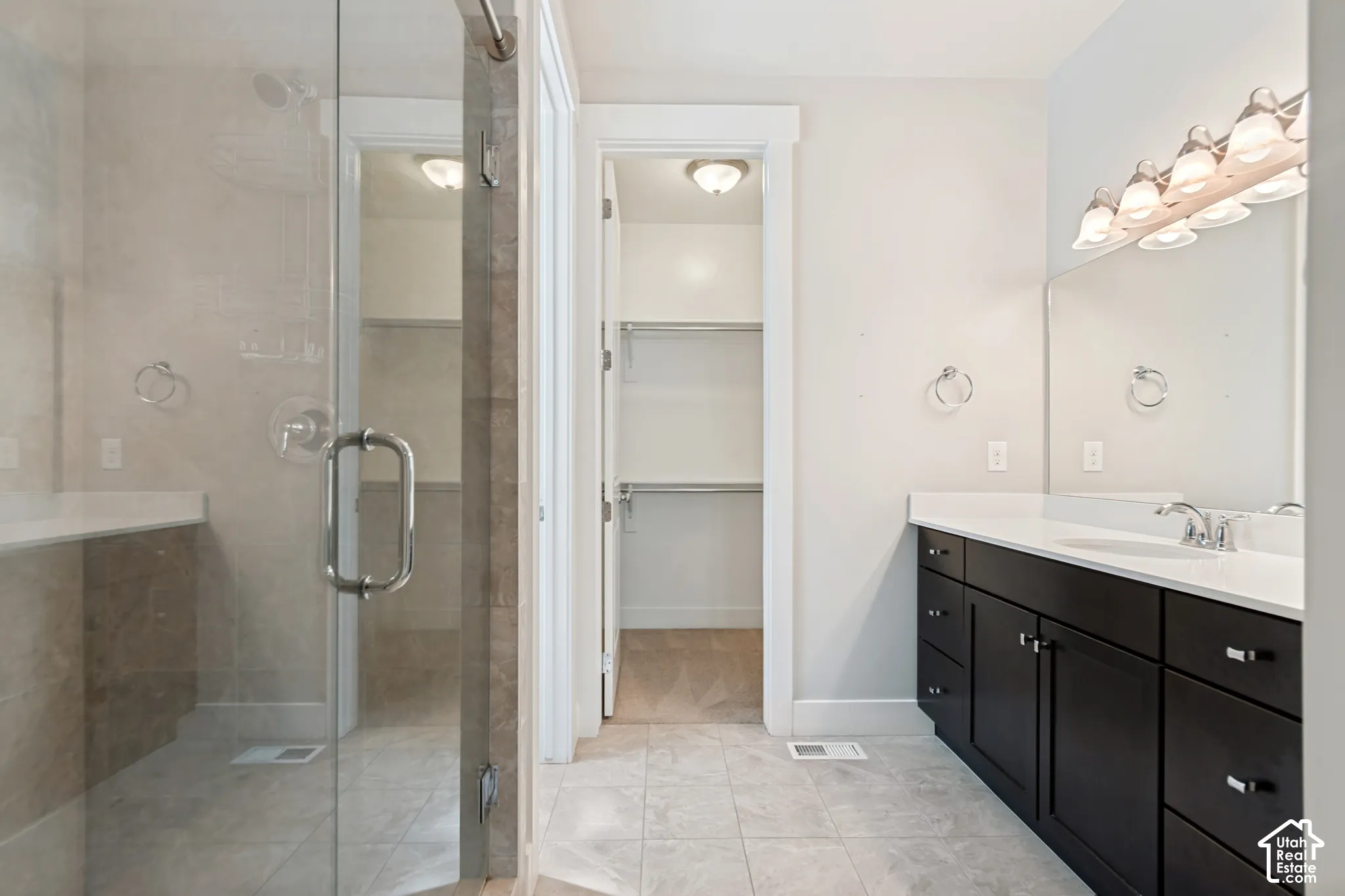 Bathroom featuring a glass-enclosed shower with tile surround, dark-finish cabinetry, solid-surface countertop, multi-light vanity fixture, and walk-in closet
