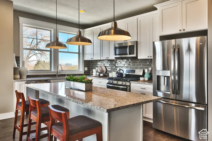 Kitchen featuring white shaker-style cabinetry, stone countertops, stainless steel appliances, a dark tile backsplash, and oversized metal pendant lighting