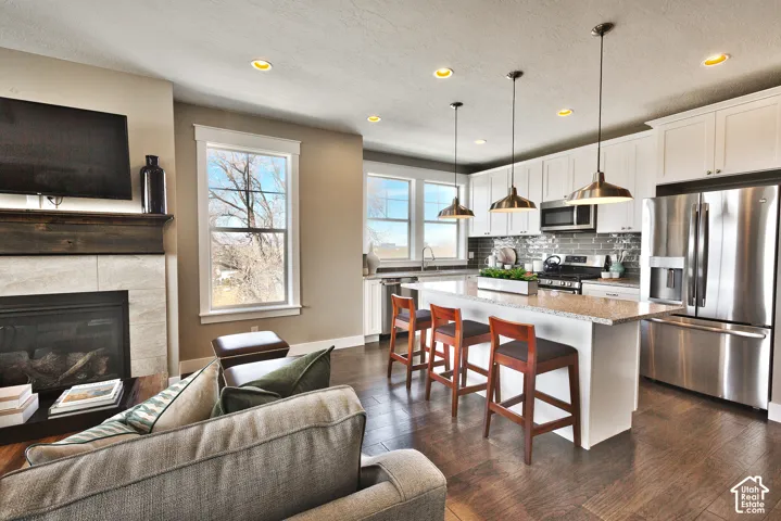 Open-concept area featuring a stone-surround fireplace with a wooden mantel, wood-finish flooring, white shaker-style cabinetry, stainless steel appliances, and a kitchen island with a stone countertop and pendant lighting