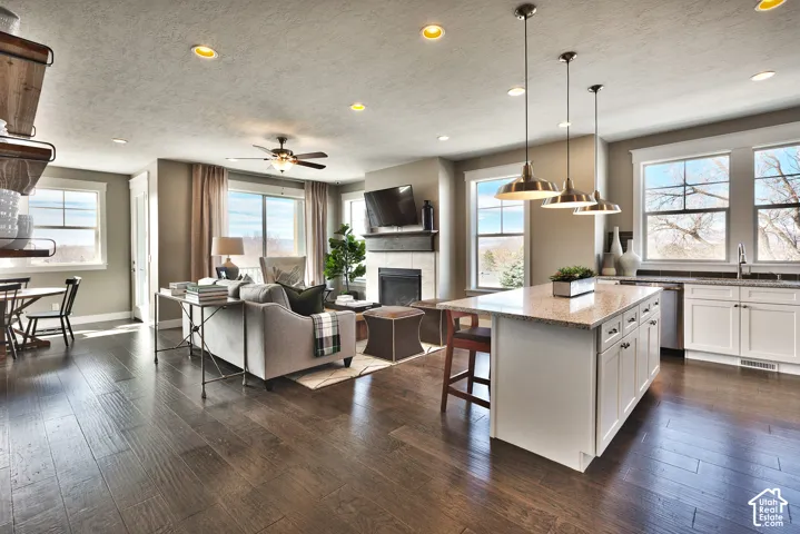 Open-concept interior featuring wood-finish flooring, a kitchen island with stone-surface countertops, recessed lighting, pendant fixtures, and a fireplace with a mantel