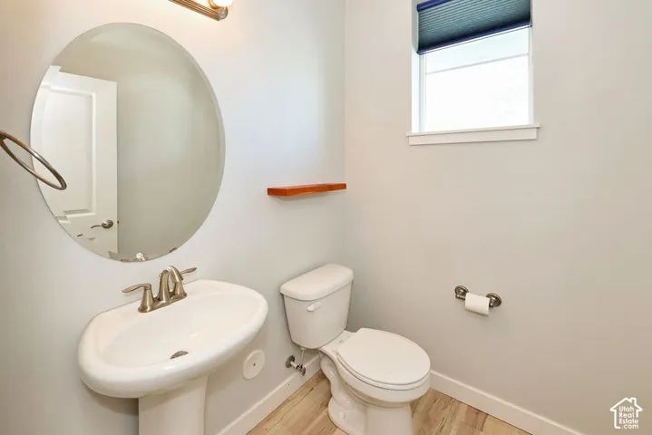 Pedestal sink with polished chrome dual-handle faucet, oval wall-mounted mirror, wood-finish flooring, and natural-wood floating display shelf