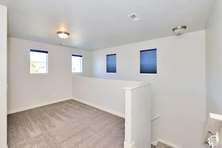 Open space featuring neutral carpeting, a partial-height pony wall, four windows with installed window coverings, and flush-mount lighting fixtures