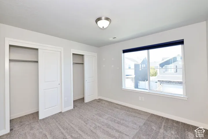 Neutral-toned interior featuring wall-to-wall carpeting, dual closets with interior shelving, a large window with a dark roller shade, and a central ceiling-mounted light fixture