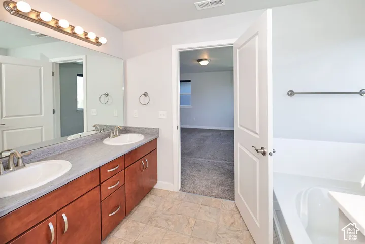 Bathroom featuring a double-sink vanity with stone-finish countertop, full-width wall mirror, wood-finish cabinetry, and a soaking tub
