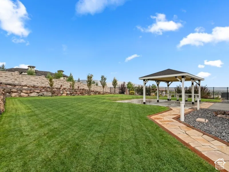Backyard featuring a manicured lawn, covered pavilion with a concrete base, flagstone pathway, tiered stone retaining wall, and metal perimeter fencing