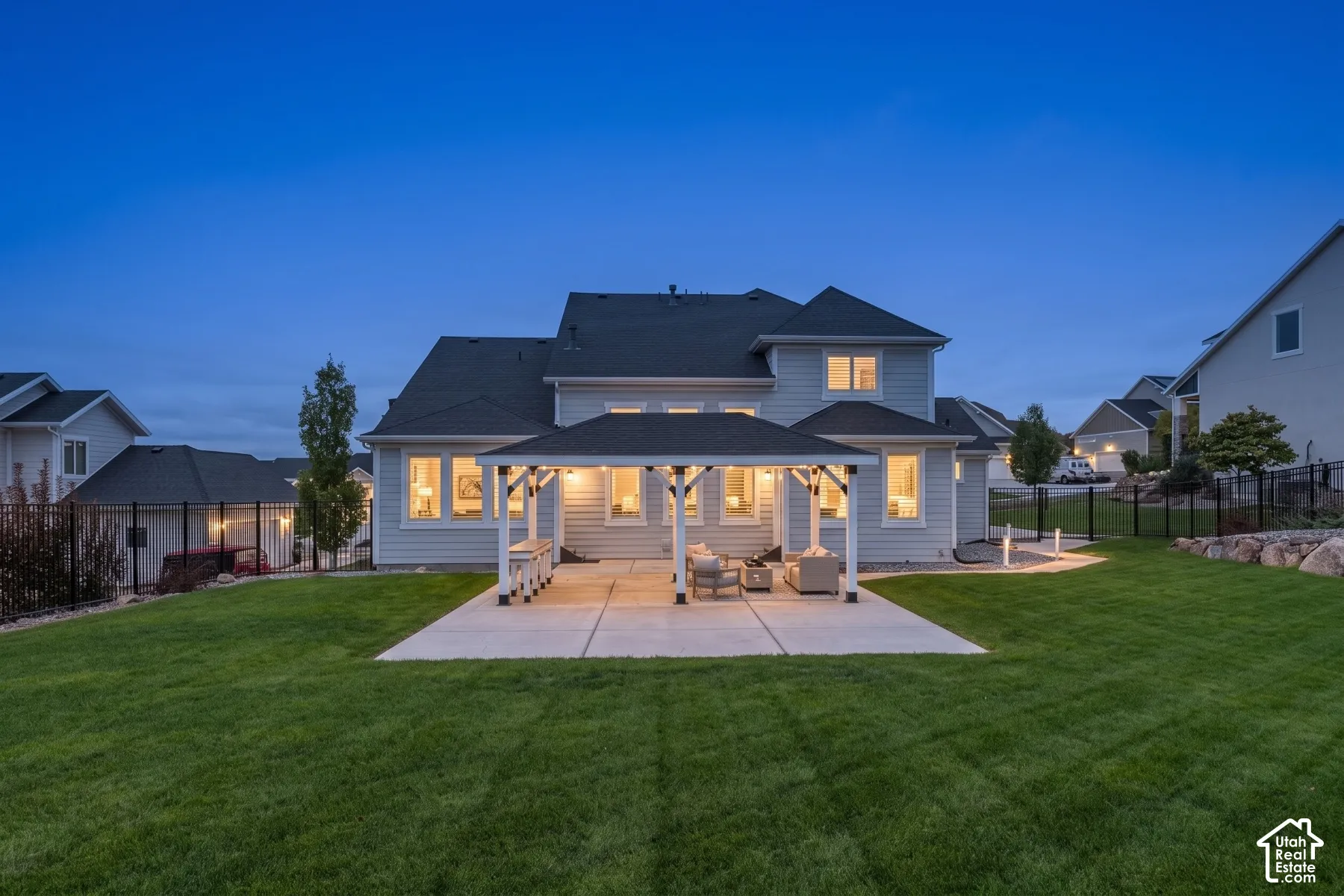 Rear exterior featuring a covered patio with a shingled roof, horizontal siding, large multi-pane windows, and a landscaped lawn enclosed by metal fencing