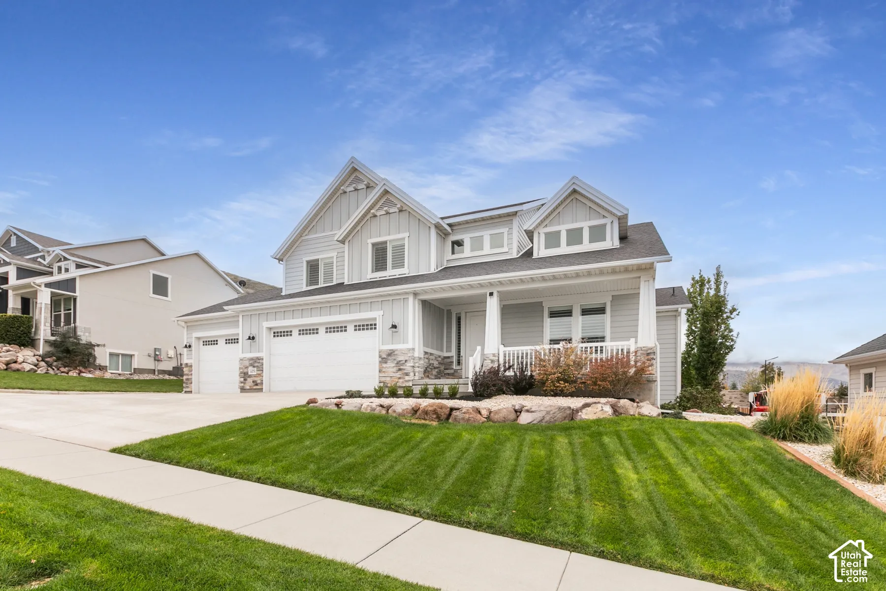 Two-story exterior featuring board-and-batten siding, stone veneer accents, an attached three-car garage, and a covered front porch with white railings