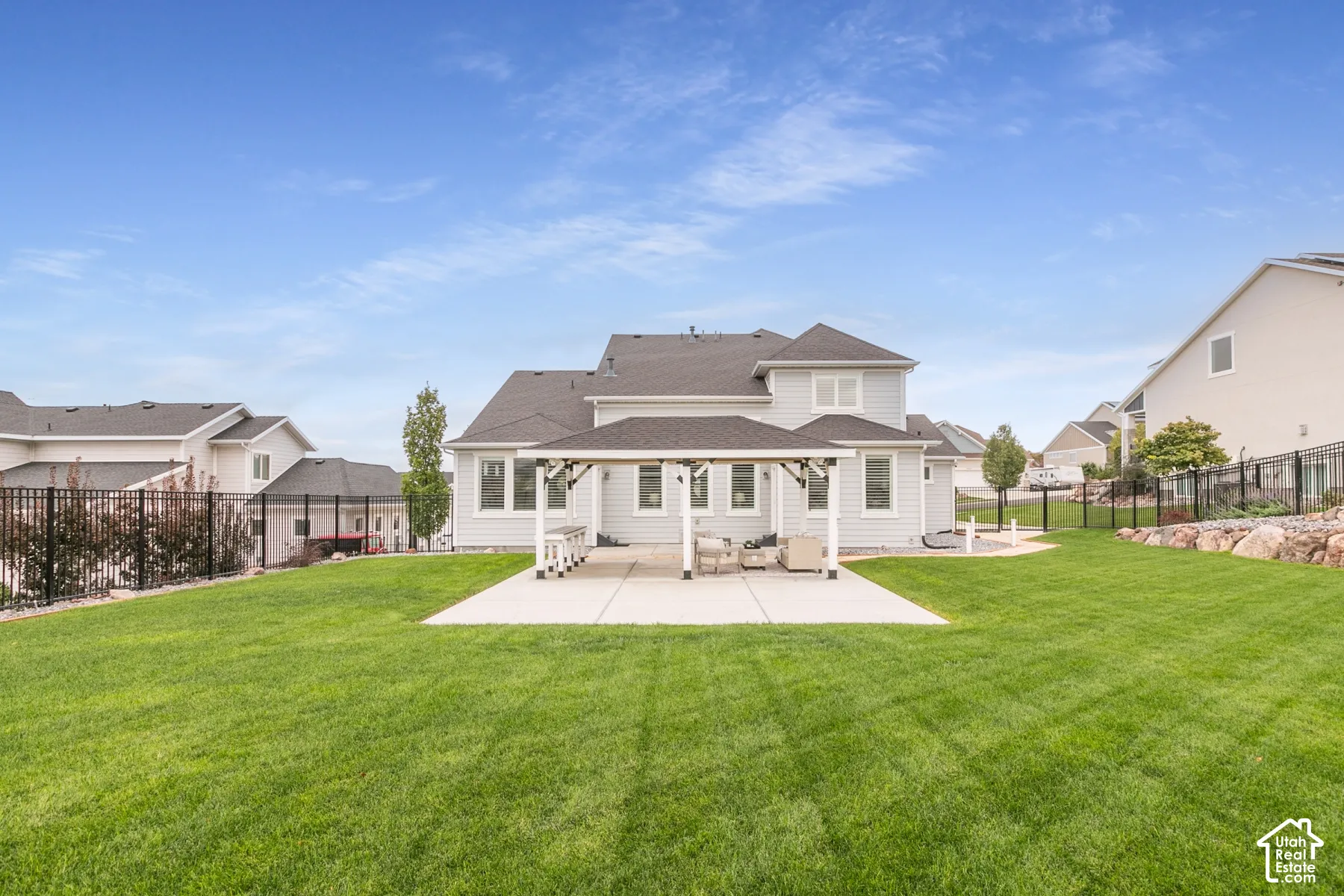 Rear exterior featuring a covered patio, concrete hardscaping, metal perimeter fencing, and a manicured lawn