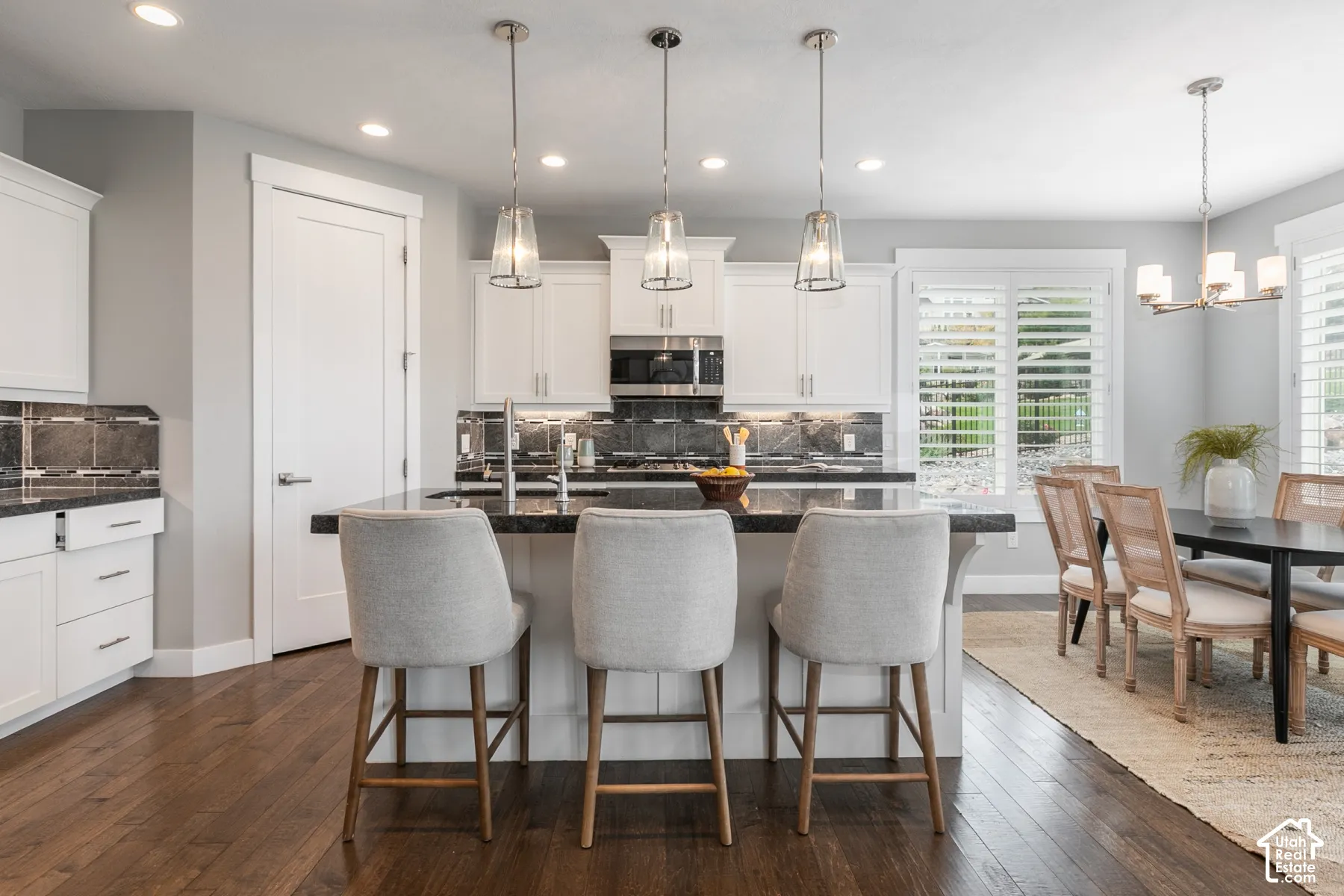 Kitchen featuring white shaker-style cabinetry, dark stone countertops, wood-finish flooring, three glass pendant lights, and plantation shutters