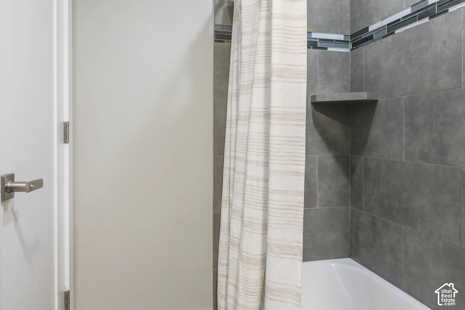 Bathtub surround featuring large-format dark grey tile, built-in corner shelving, and a glass and stone mosaic accent tile border