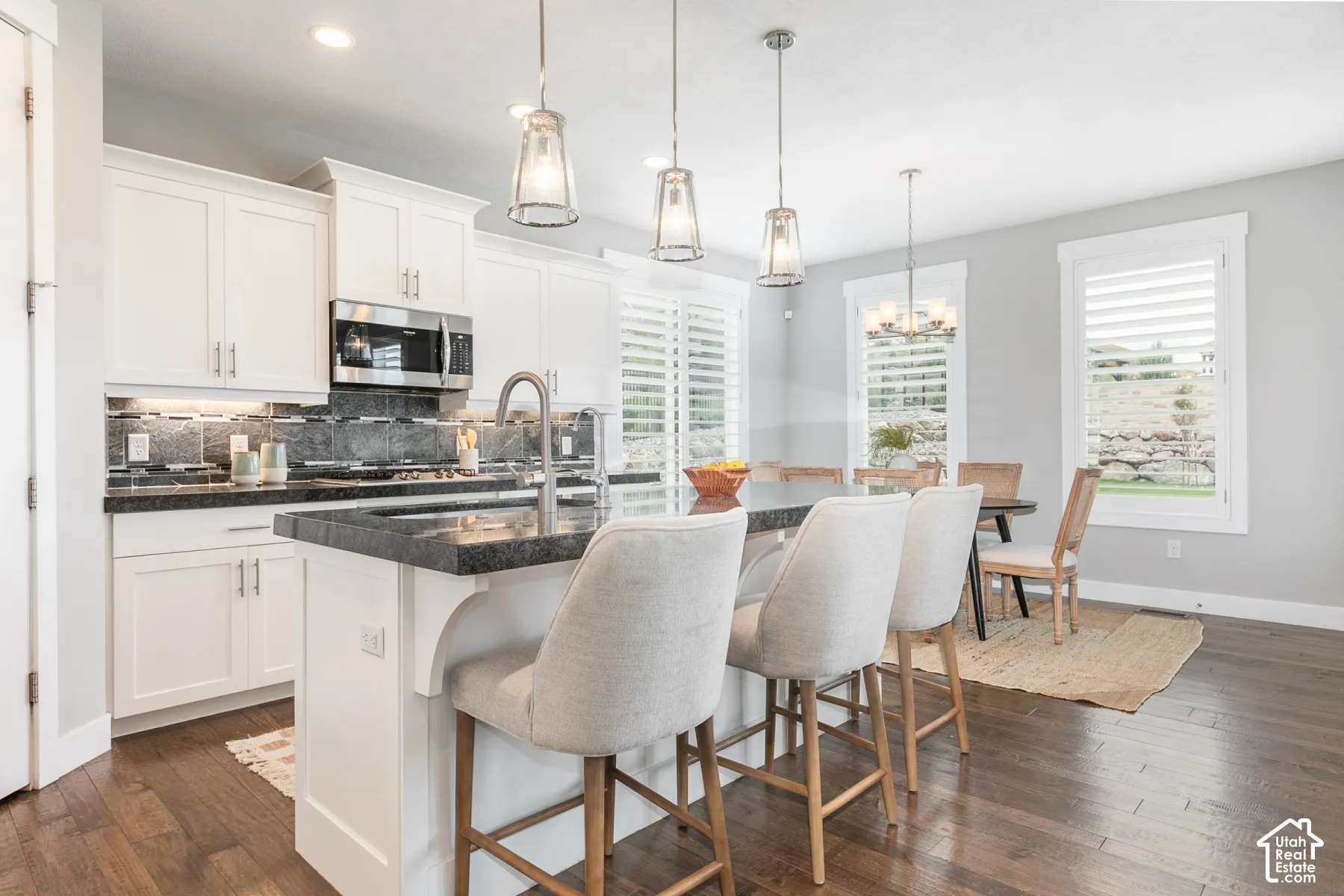 Kitchen featuring white shaker-style cabinetry, dark stone countertops, wood-finish flooring, a central island with a built-in sink, and tiered glass pendant lighting