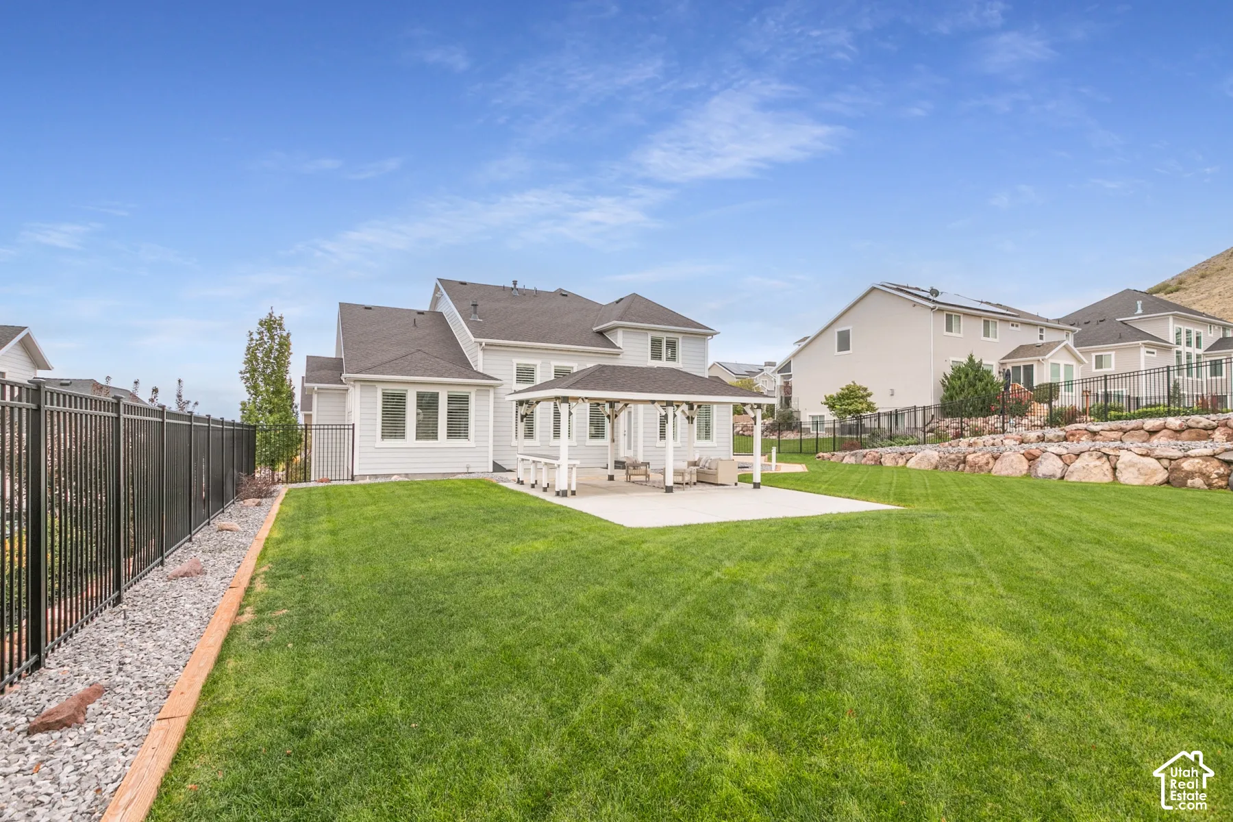 Expansive backyard featuring a covered patio, manicured lawn, metal fencing, and tiered stone retaining walls