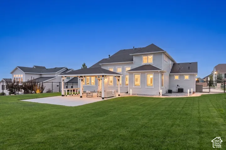 Rear exterior featuring light-toned horizontal siding, a covered patio pavilion with structural columns, an expansive lawn, and perimeter metal fencing