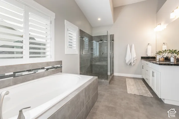 Soaking tub with tiled surround, glass-enclosed walk-in shower, white cabinetry with dark stone countertop, and shuttered windows under a vaulted ceiling