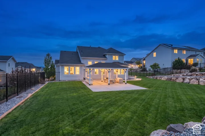 Rear exterior featuring a covered patio, tiered stone retaining walls, metal perimeter fencing, and a manicured lawn