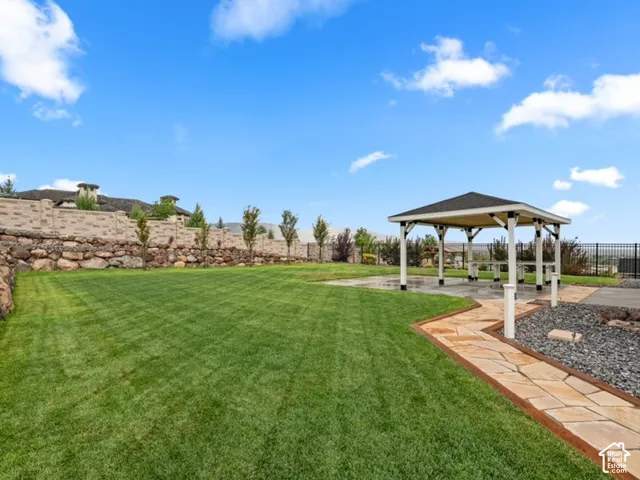 Backyard featuring a manicured lawn, covered pavilion with a concrete base, flagstone pathway, tiered stone retaining wall, and metal perimeter fencing