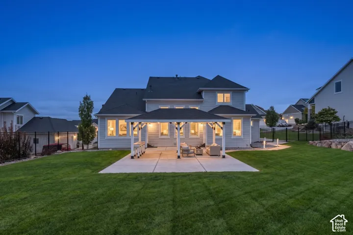 Rear exterior featuring a covered patio with a shingled roof, horizontal siding, large multi-pane windows, and a landscaped lawn enclosed by metal fencing