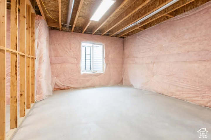 Unfinished space featuring concrete flooring, exposed wooden ceiling joists, insulated walls, a window with a metal grate, and a rectangular light fixture