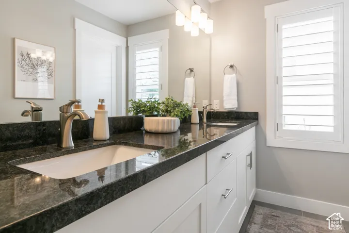 Dual-sink vanity featuring dark stone countertops, undermount rectangular basins, brushed nickel hardware, crisp white cabinetry, and plantation shutters
