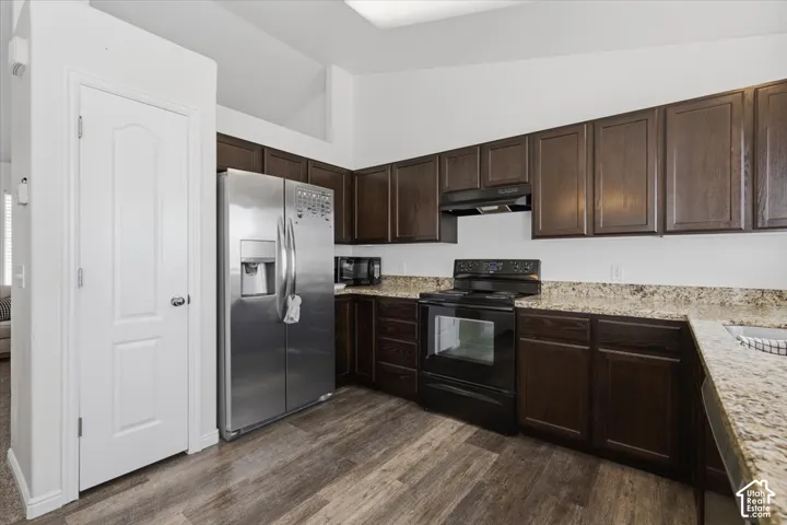Kitchen featuring dark-stained cabinetry, stone-finish countertops, stainless steel refrigerator, black range, and wood-finish flooring under a vaulted ceiling