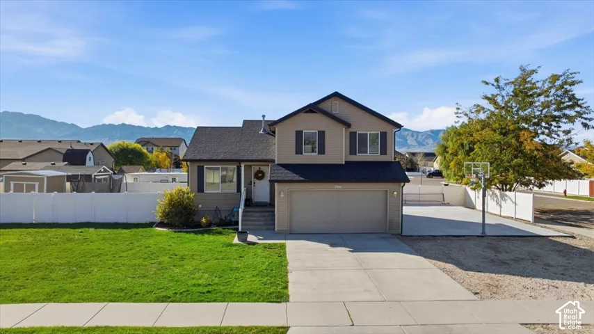 Two-story exterior featuring tan horizontal siding, an attached two-car garage, white vinyl fencing, a concrete driveway, and mountain backdrop