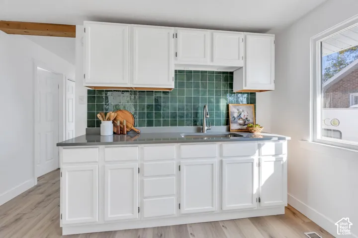 Kitchen featuring white shaker-style cabinetry, emerald green square-tile backsplash, solid-surface countertops, undermount stainless steel sink with high-arc faucet, and light wood-finish flooring