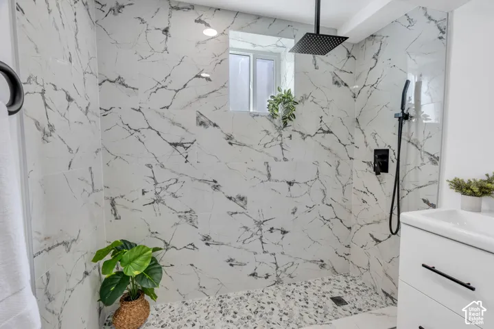 Walk-in shower featuring large-format veined tile surround, matte black rainfall shower head with handheld attachment, pebble-texture shower floor, and built-in white vanity