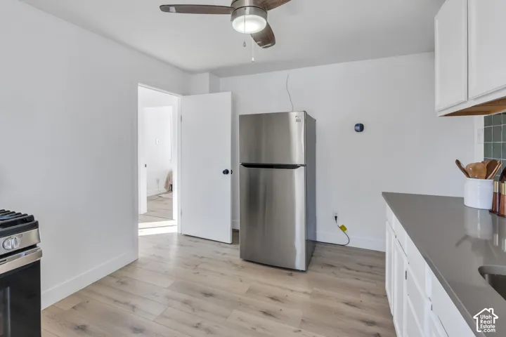 Kitchen area featuring wood-finish flooring, stainless steel refrigerator, matte grey solid-surface countertop, white shaker-style cabinetry, and a ceiling fan with integrated lighting