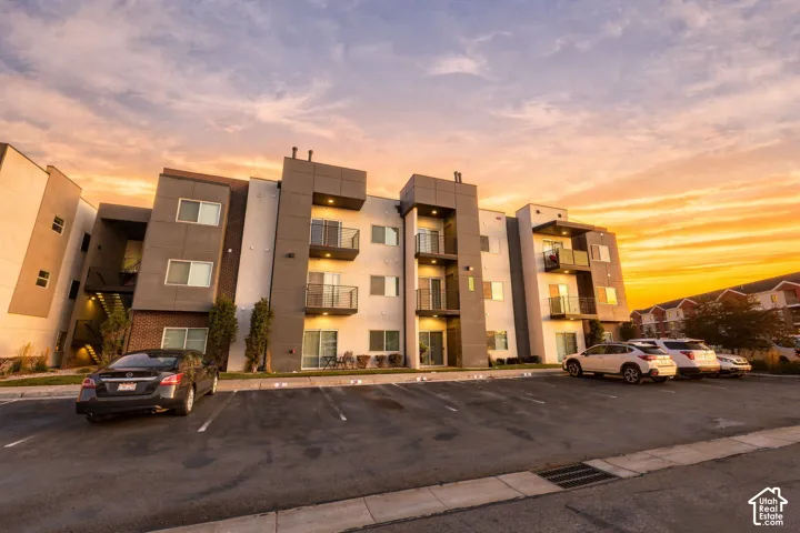 Modern multi-story exterior featuring a grey and white stucco facade, brick accents, integrated balconies with metal railings, and asphalt parking area