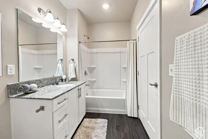 Bathroom featuring white cabinetry, stone-finish countertop, undermount sink, wood-finish flooring, and a shower-tub combination with integrated shelving