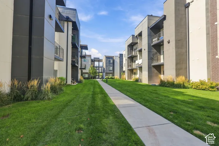 Modern multi-story exterior featuring metal paneling, private balconies, a concrete pedestrian walkway, and manicured green space with ornamental grasses