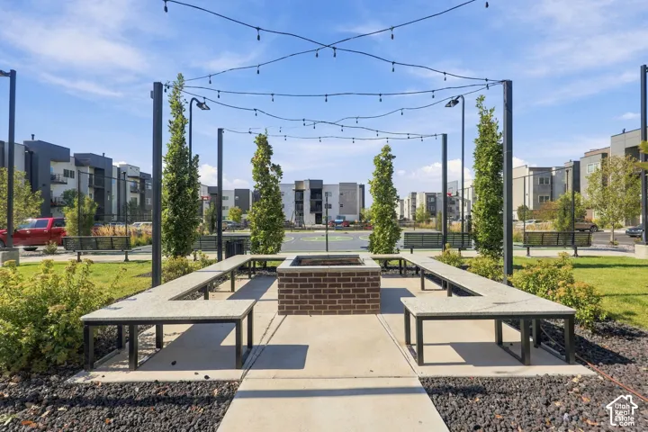 Outdoor common area featuring a central brick fire pit, integrated stone-topped bench seating, overhead string lighting, and ornamental landscaping