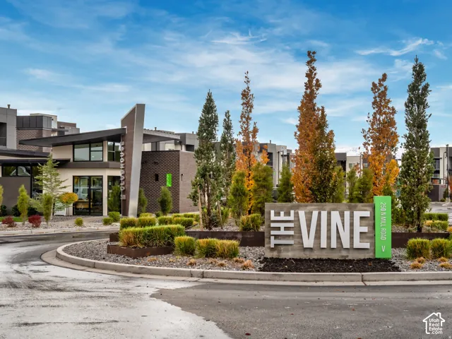 Community entrance featuring a concrete monument sign, mature evergreen and deciduous landscaping, gravel hardscaping, and a contemporary clubhouse building with a multi-level roofline