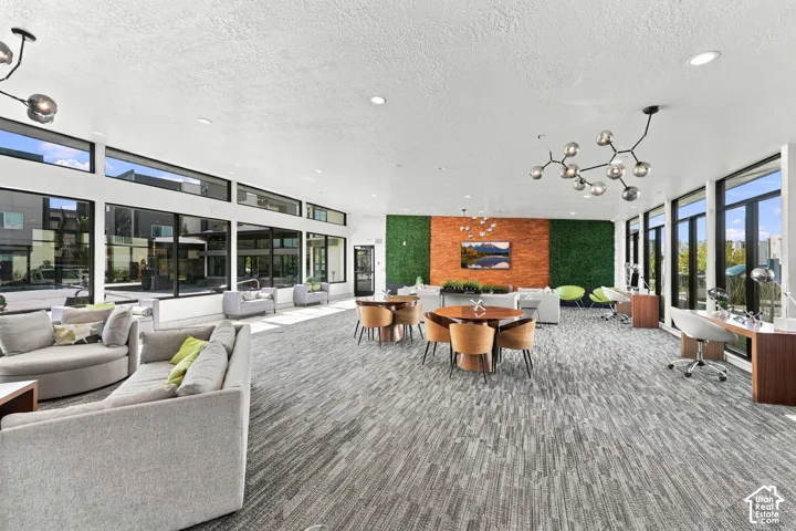 Expansive open-concept clubhouose featuring floor-to-ceiling windows, patterned carpeting, recessed lighting, architectural multi-bulb chandeliers, and a decorative feature wall with integrated greenery and textured stone cladding