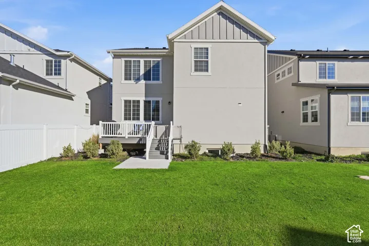 Rear exterior featuring a multi-level deck with white railings, a concrete patio, landscaped beds with shrubbery, and vinyl privacy fencing surrounding a grass lawn
