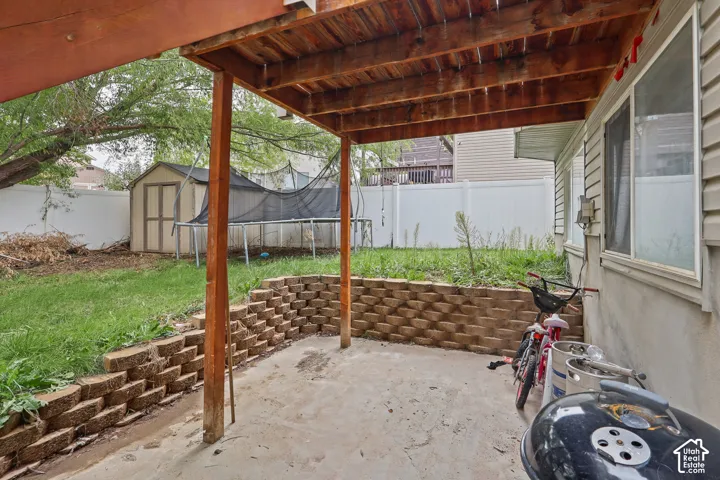 Covered concrete patio featuring a timber support structure, segmented masonry retaining wall, and fenced backyard with a detached storage shed