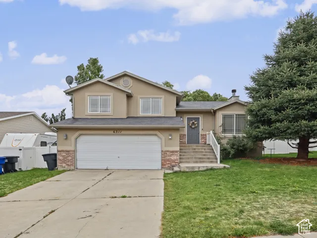 Split-level exterior featuring stucco siding, stone accents, a concrete driveway, a two-car garage, and a mature evergreen tree