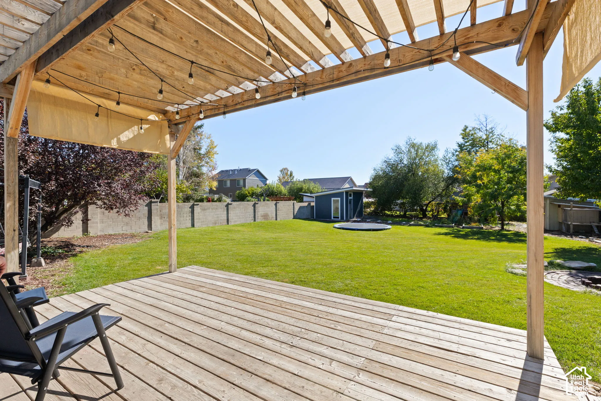 Wood-plank deck featuring a wooden pergola with overhead string lighting, expansive grass yard, perimeter concrete masonry wall, and detached storage structure