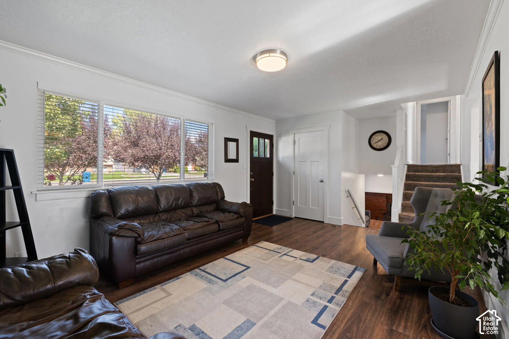 Wood-finish flooring, large picture window with horizontal blinds, flush-mount ceiling lighting, multi-panel front door, and stairwell with carpeted treads