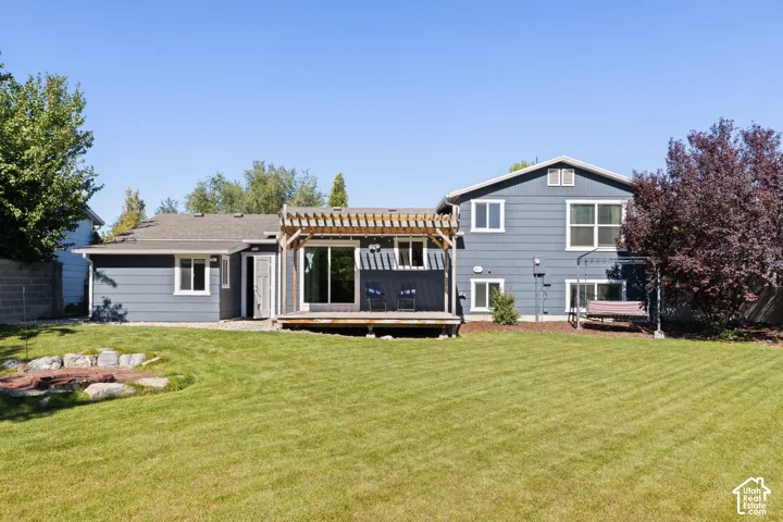 Rear exterior featuring blue horizontal siding, a wood pergola, a wood deck, and a stone-bordered fire pit within a landscaped lawn