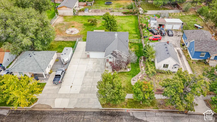 Aerial perspective of a single-story residence featuring a paved driveway, shingled roofing, mature landscaping, and an expansive rear lawn