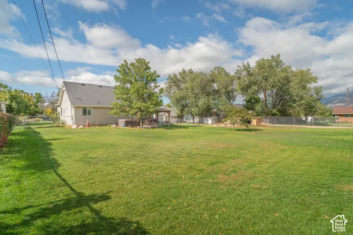 Expansive backyard featuring a manicured lawn, mature trees, chain-link perimeter fencing, and a covered patio area