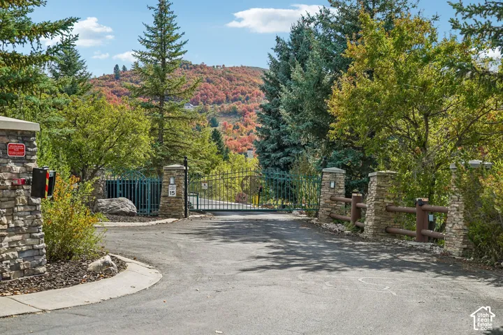 Stone-clad entry pillars, decorative metal gate, and asphalt driveway framed by mature trees and natural landscaping