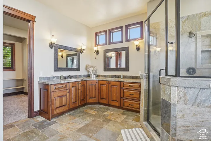 Dual wood-finish vanity with granite countertops and undermount sinks, natural stone tile flooring, and a glass-enclosed shower featuring natural stone tiling