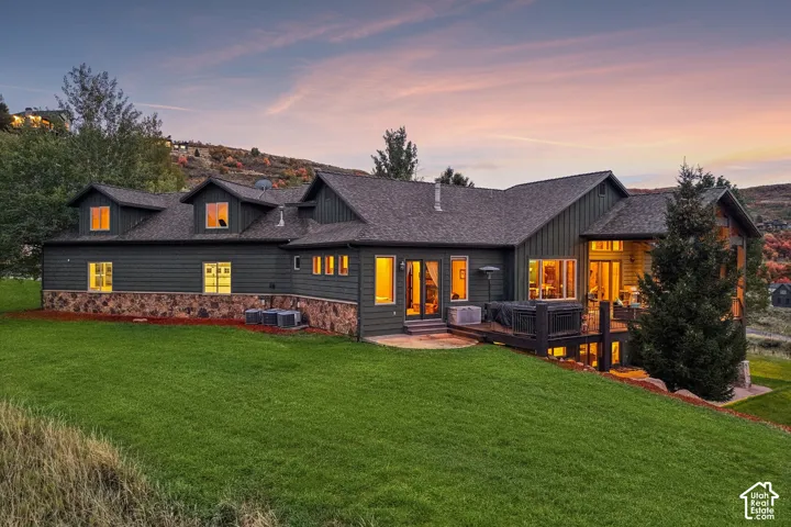 Expansive rear facade featuring dark siding, stone accents, multiple dormer windows, a spacious wood deck, and a stone patio